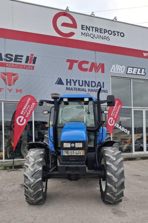 Trator Convencional New Holland TS115 em frente a um prédio com a inscrição 'Entreposto e Máquinas" e várias marcas. Mostra também uma bandeira vermelha com a inscrição "Entreposto Máquinas usados' indicam a venda de usados.